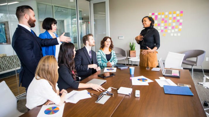 An agile project management meeting in a room with a large table and a group of employees on one side of the desk and one woman on the other side of the table giving a presentation -- behind her is a wall and a cluster of post it notes stuck to the wall.