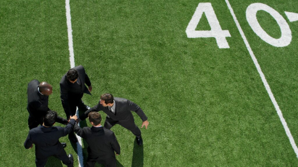 A team of businessmen in suits and ties on a football field in a huddle to represent teamwork.