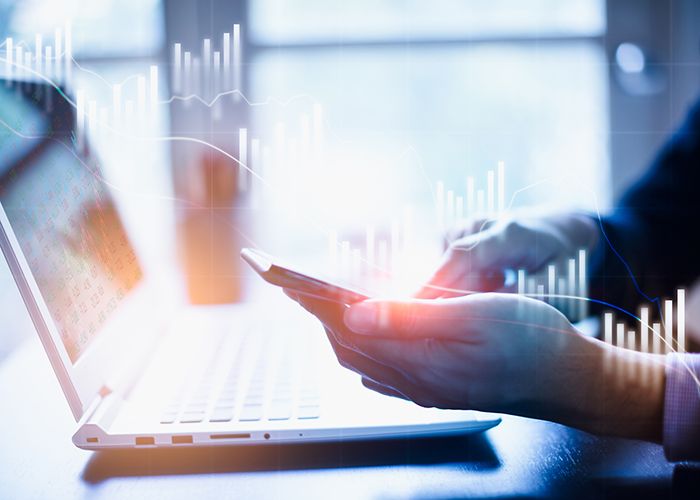 A pair of hands using a smartphone over a laptop that has holographic line charts floating around it.