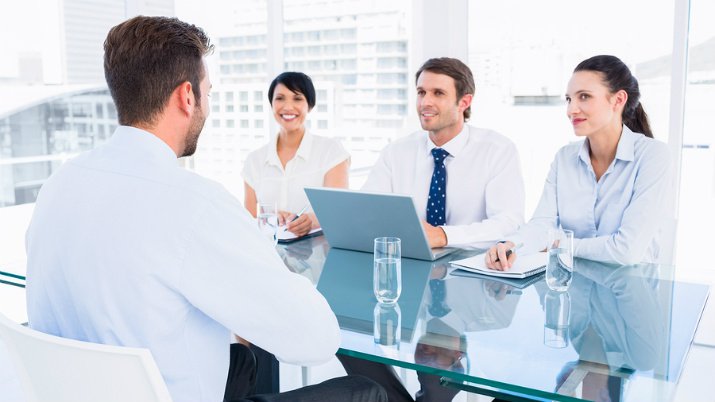 Three people sitting on one side of a glass table interviewing a man sitting across from them.