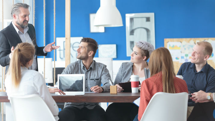 A modern workspace with vibrant blue walls and a group of co-workers sitting at a table looking up at a man standing up giving them instructions.