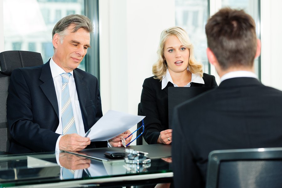 Three business people sitting around a table, with one man on one side of a table and a man and a woman sitting across from him looking at a piece of paper and talking to him as if they are interviewing him.