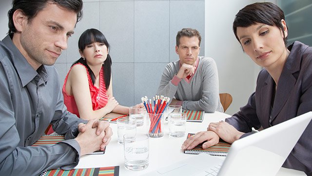 A group of four people working together at a messy table with drinks, papers, pens, and notebooks while looking at a laptop at the end of a table.