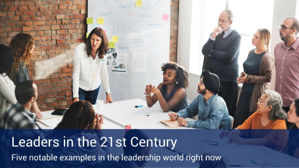 An office full of people working together talking to each other around a table with a large whiteboard on the wall that says "Ideas" in marker.