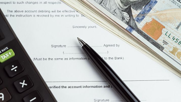 Close up of a contract with a pen sitting on top of the signature line, with a stack of hundred dollar bills and a calculator sitting on top of the contract.