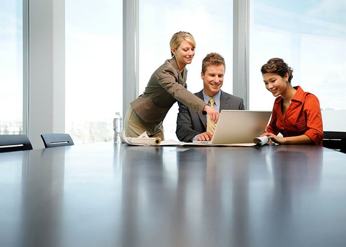 Three people in a highrise office at a board table all looking at a laptop with a window behind them overlooking a city.