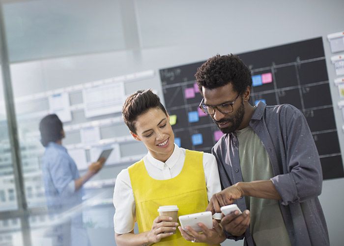 A woman wearing a yellow vest is holding a tablet and a cup of coffee while helping a project management coworker with scheduling on his smartphone.