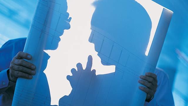 A silhouette of two men looking at a large piece of rolled out paper with blueprints on it.