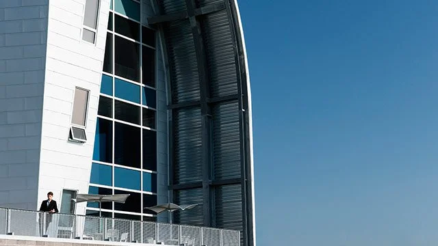 A large modern building with big windows and a man standing on the balcony looking out over the city.